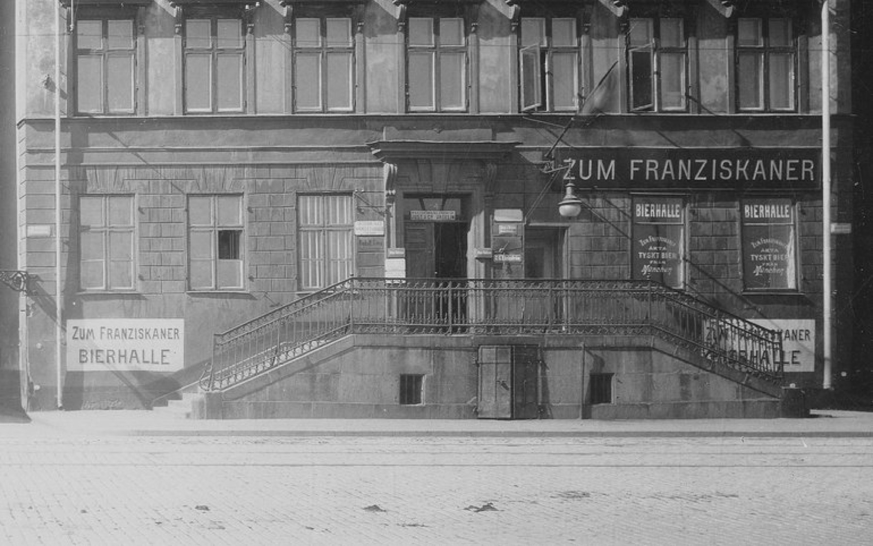 Gammalt svartvitt foto av fasaden mot Skeppsbron, med skyltarna Zum Franziskaner och Bierhalle över dubbeltrappan