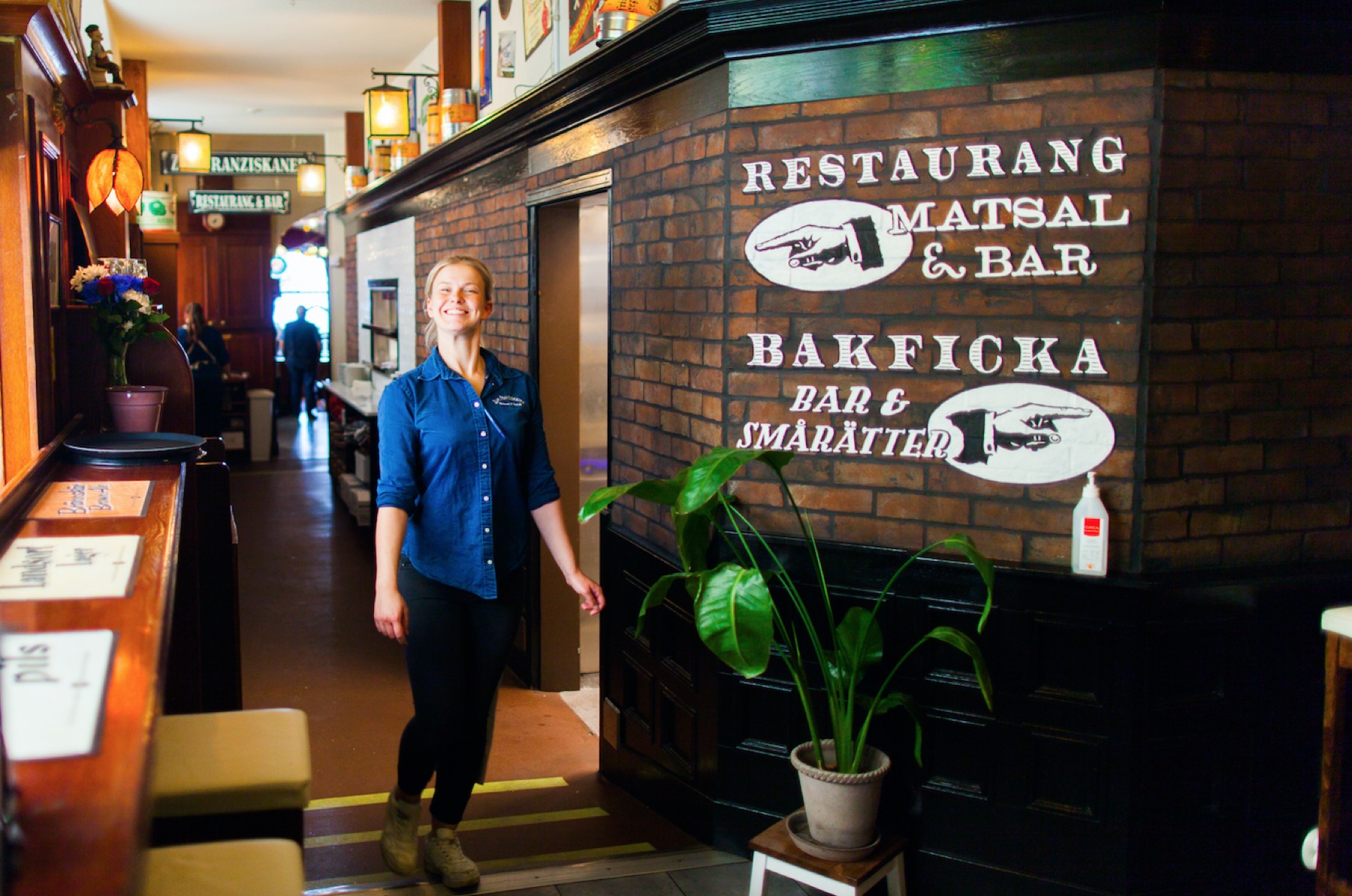 Medarbetare i Zum Franziskaner-skjorta i entrén, vid de handmålade skyltarna Restaurang matsal och bar och Bakficka bar och smårätter