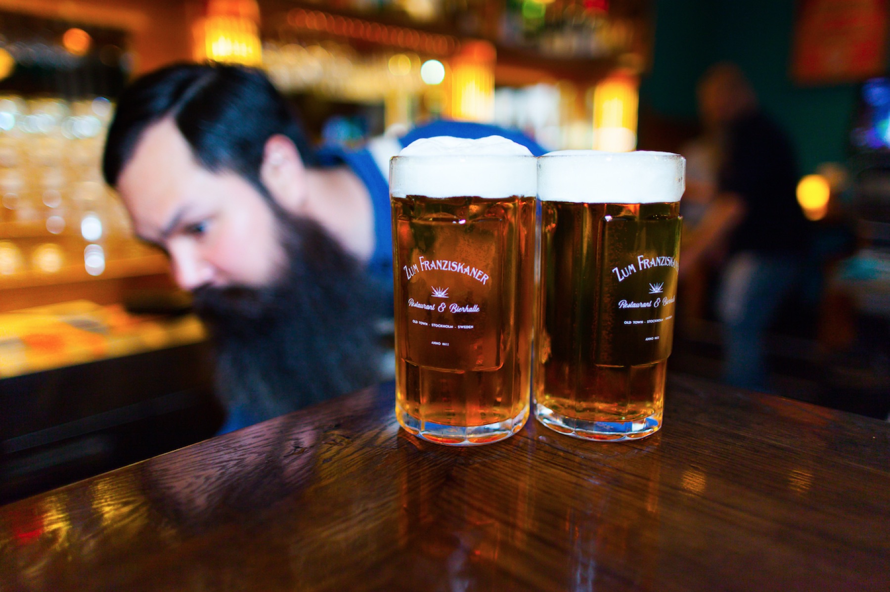 Två fyllda Zum Franziskaner-glas på bardisken med en långskäggig bartender i bakgrunden
