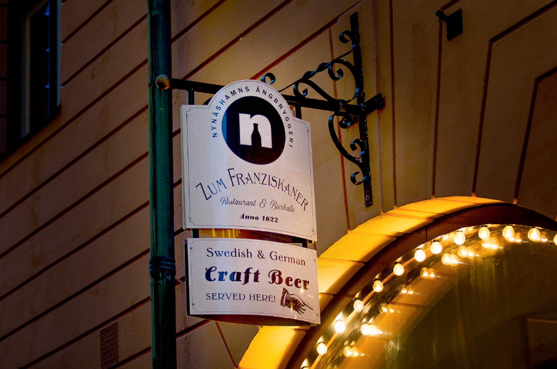 Skylten Zum Franziskaner Restaurant och Bierhalle på fasaden om kvällen, intill den ljusslingade valvportalen
